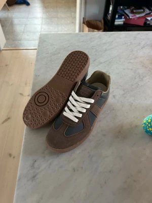 Maison margiela Gats  - Snygga sneakers i brunt och grått med vita skosnören. Skorna har en klassisk låg profil och är tillverkade i en mix av mocka och slätt läder. Yttersulan är i gummi och ger bra grepp. Perfekta för dig som gillar stilrena och tidlösa sneakers. 