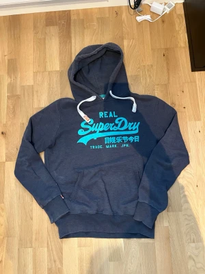 Mörkblå Superdry hoodie med tryck - Snygg mörkblå hoodie från Superdry med turkost tryck på bröstet och vita dragsnören i huvan. Klassisk känguruficka framtill och ribbade muddar vid ärmar och nederkant. Perfekt för en avslappnad och cool stil. Trycket ser lite slitet ut men fortfarande ett väldigt snyggt plagg! Passar M-L