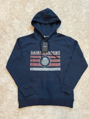 Mörkblå Saint Laurent hoodie - Snygg mörkblå hoodie från Saint Laurent med tryck i vitt och rött framtill. Klassisk huva med dragsko och känguruficka. Trycket har en vintage-look med texten SAINT LAURENT och ett emblem med SL i mitten. Perfekt för en avslappnad streetwear-stil. ⚠️‼️tröjan är i storlek M men passar bättre S‼️⚠️