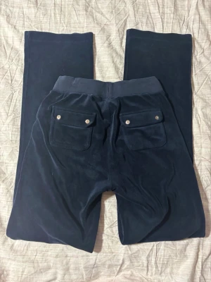juicy byxor  - Mörkblå mjukisbyxor i mjuk velour med resår och dragsko i midjan. Byxorna har två fickor fram och två bakfickor med lock och silverfärgade knappar. Perfekta för en chill och avslappnad stil.