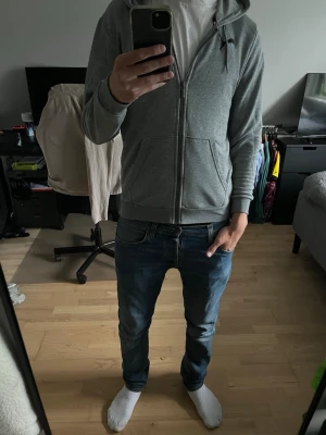 Grå zip hoodie med huva från Puma - Säljer en stilren grå zip hoodie från Puma med huva och två stora fickor framtill. Tröjan har ribbade muddar vid ärmslut och nederkant samt Puma-logga på bröstet. Perfekt för en avslappnad och sportig look.