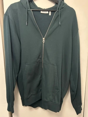 Mörkgrön hoodie med dragkedja Weekday - Mörkgrön hoodie från Weekday med dragkedja framtill, justerbar huva med snören och stor känguruficka. Tillverkad i mjukt bomullsmaterial och har ribbade muddar vid ärmslut och nederkant. Perfekt för en avslappnad och stilren look.