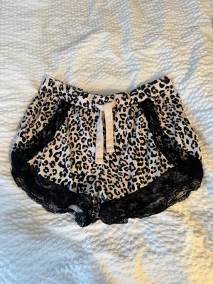 Leopardmönstrade pyjamasshorts med spets - Snygga pyjamasshorts från New Woman i leopardmönster med svarta spetsdetaljer längs kanterna och en ljus rosett framtill. Perfekta för mysiga hemmakvällar. Mjukt och bekvämt material.