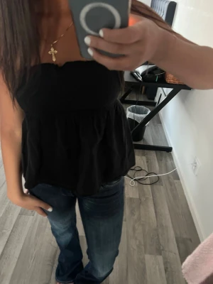 Svart axelbandslös tubtopp - Säljer en svart, axelbandslös tubtopp med volang nedtill. Toppen har en bandeauformad överdel och sitter snyggt över bysten. Perfekt att matcha med jeans eller kjol för en trendig look.