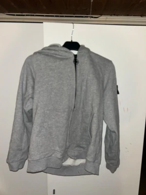 Grå hoodie från Stone Island - Grå zip-hoodie från Stone Island med huva och dragkedja framtill. Klassisk modell med ribbade muddar och fickor på sidorna. Tillverkad i mjukt bomullsmaterial och har den ikoniska loggan på ärmen.