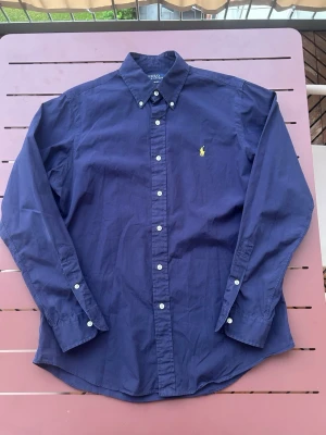 Polo Ralph Lauren skjorta - Riktigt snygg Polo Ralph Lauren skjorta i storlek M. Inga defekter som jag har märkt. Fin marinblå färg med en gul polo logga. Riktigt skön och funkar perfekt nu under sommaren. Pris går att diskutera vid snabb affär. Skickar inom 24h.