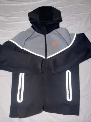 Nike Tech Fleece hoodie svart/grå - Nike Tech Fleece hoodie i svart och grått med reflekterande detaljer och orange Nike-logga på bröstet. Dragkedja framtill, två fickor med dragkedja och tumhål i ärmsluten. Tillverkad i mjukt och bekvämt material. Perfekt för en sportig och stilren look.