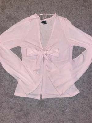 Ljusrosa omlottopp med rosett - Supersöt ljusrosa omlottopp med lång ärm och stor rosett framtill. Toppen är gjord i ett tunt, mjukt material och har en feminin, croppad passform. Perfekt att styla med jeans eller kjol för en trendig look.
