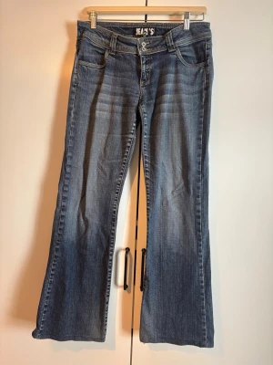 Blå bootcut jeans med slitningar - Säljer ett par blå bootcut jeans med snygga slitningar och ljusare partier framtill. Jeansen har fem fickor, bälteshällor och dubbla knappar i midjan. Bakfickorna har dekorativa detaljer. Perfekta för en avslappnad och trendig look.