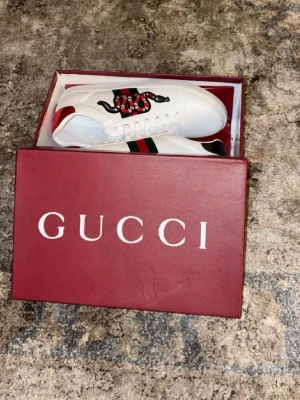Gucci vita sneakers med orm - Snygga vita sneakers från Gucci med broderad röd och svart orm på sidan samt klassiska gröna och röda ränder. Baksidan har metallicdetaljer i rött och grönt med Gucci-logga. Skorna är i läder och har en stilren, låg profil. Skorna e knappast använda och e i fint skick priset inte hugget i sten direkt , hör av er om ni e sugna 💯