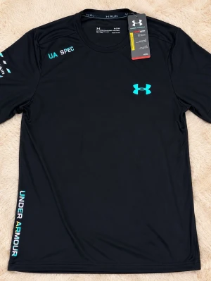 Svart tränings-tshirt Under Armour - Snygg svart tränings-tshirt från Under Armour med turkosa loggor och textdetaljer. Tillverkad i ett lätt och snabbtorkande funktionsmaterial som passar perfekt till löpning eller gym. Rund halsringning och korta ärmar för maximal rörelsefrihet. Storlek M och S