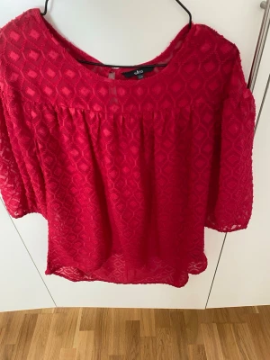 Röd transparent blus - Snygg röd blus från ellos  i transparent material med diskret mönster. Blusen har rund halsringning och trekvartsärm. Perfekt att styla med ett linne under för en trendig look.