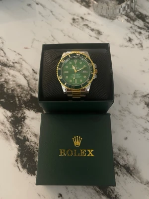 Rolx Submariner grön/guld klocka - Snygg Rolx Submariner med grön urtavla och gulddetaljer. Klockan har en tvåfärgad länk i silver och guld samt tydliga indexmarkeringar. Kommer i box. Perfekt accessoar för dig som vill sticka ut med stil.