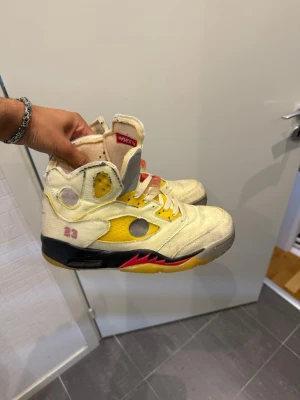 Off-White x Air Jordan 5 gul/svart - Säljer ett par Off-White x Air Jordan 5 sneakers i gul mesh och läder med svarta och röda detaljer. Skorna har högt skaft, genomskinliga paneler, gula skosnören och tydliga Off-White detaljer. Sulan är gul med röda och svarta inslag samt ikoniska Jumpman-loggan undertill.
