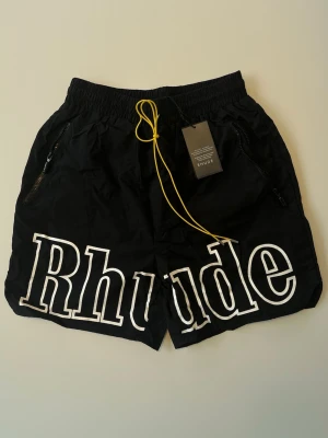 Rhude Shorts - Svart - Rhude shorts i svart nylon med stort vitt “Rhude” tryck fram, gul dragsko-detalj i kontrast. Dragkedjefickor på båda sidor, avslappnad passform perfekt för sommaren.  Skick: Nyskick, prislapp kvar  Storlek: L  Skickas snabbt och säkert! 📦