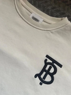 Vit sweatshirt från Burberry - Vit sweatshirt från Burberry med klassisk rund halsringning och svart broderad TB-logga på bröstet. Tillverkad i mjukt material och har ribbade muddar vid ärmslut och nederkant. Perfekt för en clean och stilren look.  Jag säljer endast denna för att den börjar bli för liten. Pris går att diskuteras!