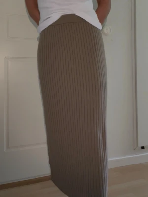Stickad kjol - Säljer en ribbad maxikjol i beige/brun färg. Kjolen har en rak, lång passform och bred resår i midjan för skön komfort. Materialet är mjukt och stretchigt, perfekt för en avslappnad men ändå stilren look.