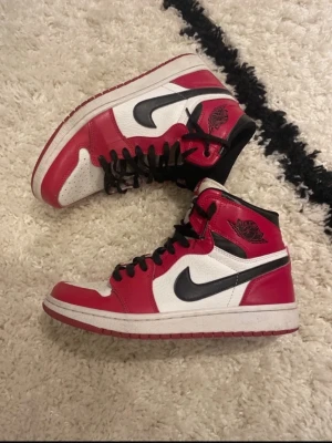 Nike Air Jordan 1 High röd/vit - Säljer ett par Nike Air Jordan 1 High sneakers i rött, vitt och svart. Skorna har klassisk hög siluett, svart swoosh på sidan, perforerad tåbox och svarta skosnören. Tillverkade i läder med ikoniska Air Jordan-loggan på sidan. Storlek 43