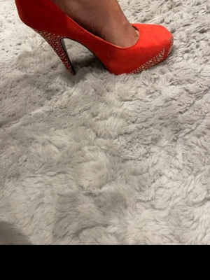 Röda pumps med strassdetaljer - Snygga röda pumps med hög klack och öppen tå. Skorna har strassdetaljer framtill och på klacken som ger extra bling. Materialet ser ut att vara mockaimitation och färgen är riktigt klar röd. Perfekta för dig som vill sticka ut.