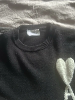 Svart stickad tröja med hjärtdetalj - Svart stickad tröja med rund halsringning och ribbade muddar. På bröstet finns en stor vit hjärtformad detalj med ett A under, vilket ger en unik och trendig look. Perfekt för dig som gillar stilrena men ändå lekfulla plagg.