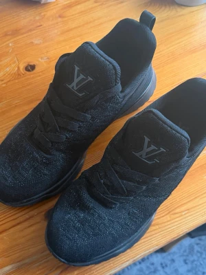 Svarta Louis Vuitton sneakers - Snygga svarta sneakers från Louis Vuitton med stickad ovandel och klassisk LV-logga på plösen. Skorna har svarta skosnören, diskreta detaljer på hälen och en robust sula med LV-symboler. Perfekta för en trendig och stilren look.