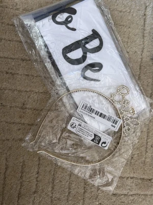Guldigt diadem med Bride-text - Snyggt diadem i guldfärgad metall med ordet 'Bride' dekorerat med glittrande stenar. Perfekt accessoar till möhippa eller bröllopsfest för att verkligen sticka ut. Enkel och stilren design som ger en festlig känsla. Bride to be band ingår också 