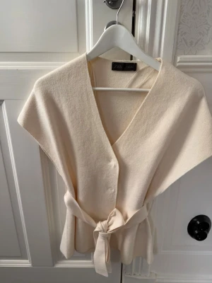 Beige omlottväst med bälte från Zara - Snygg beige väst från Zara i omlottmodell med knytbälte i midjan. Västen har v-ringad hals och är tillverkad i ett mjukt material som ger en avslappnad men stilren look. Perfekt att slänga över en tröja för extra lager.