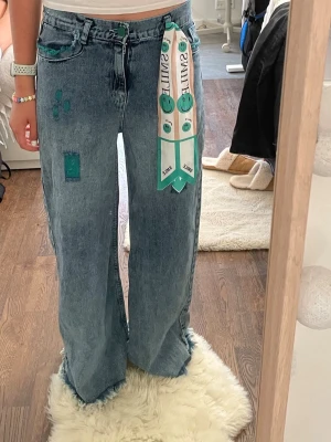 Baggy jeans med patchar och scarf - Säljer ett par ljusblå baggy jeans med gröna patchar och råa kanter nertill. Jeansen har en cool scarf med smileys och texten 'SMILE' i midjan. Femficksmodell och hög midja. Perfekta för dig som gillar en avslappnad och unik stil.