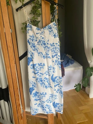 Vit och blå blommig sommarklänning - Säljer en vit långklänning med blått blommönster och tunna axelband. Klänningen har en v-ringad urringning och är perfekt för varma sommardagar. Har en liten fläck (se bild) men rätt säker på att den går bort.