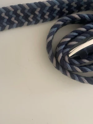 Flätat blått och grått bälte - Säljer ett snyggt flätat bälte i olika nyanser av blått och grått. Bältet har ett coolt mönster och ett silvrigt spänne som ger en modern touch. Perfekt för att lyfta vilken outfit som helst.