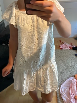 Vit kort klänning med volang - Söt vit kort klänning med broderade hål och volang nertill. Klänningen har korta ärmar och fyrkantig ringning, perfekt för varma dagar. Tillverkad i bomull för en luftig och bekväm känsla.