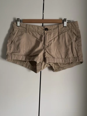 Beige shorts från So - Säljer ett par klassiska beige shorts från So med normal passform. De har fickor fram, bälteshällor och stängs med knapp och dragkedja. Perfekta för varma dagar och enkla att matcha med olika toppar.