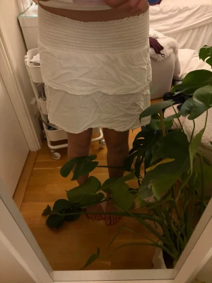 Vit kortkjol med smock i midjan - Säljer en vit kortkjol med smock i midjan och volangdetaljer. Kjolen har en luftig och bekväm passform, perfekt för varmare dagar. Enkel att matcha med olika toppar för en fräsch look.