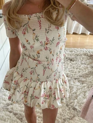 Blommig offshoulder kortklänning - Supersöt kortklänning med offshoulder och volangkant. Klänningen är vit med blommigt mönster i rosa, gult och grönt. Smockad överdel ger en bekväm passform och kjolen har volanger för extra flow. Perfekt för varma dagar och sommarvibbar!