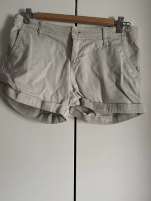 Beige shorts med uppvikta ben - Säljer ett par ljusbeige shorts med uppvikta ben och klassisk knapp- och dragkedjestängning. Shortsen har sidofickor fram och passpoalfickor bak. Perfekta för varma dagar och har en enkel, stilren look.
