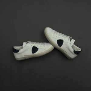 Vita Philippe Model sneakers med blåa detaljer - Snygga vita sneakers från Philippe Model med blåa detaljer på sidan och hälen. Skorna har klassisk låg profil, vita skosnören och en vit sula. Tillverkade i läder för en clean och stilren look som passar till det mesta.