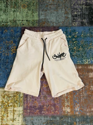 Bershka shorts - Säljer ett par beiga shorts med svart tryck på vänster ben och svart snörning i midjan. Shortsen har elastisk midja, sidofickor och en bakficka. Perfekta för sommaren och tillverkade i mjuk bomull.