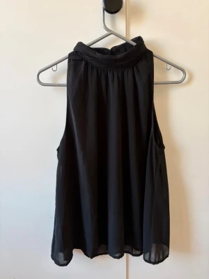 Svart ärmlös blus med hög krage - Säljer en svart, luftig blus med hög krage och ärmlös design. Toppen har ett lätt transparent material och ett snyggt fall. Perfekt för dig som gillar stilrena och enkla plagg med en twist.