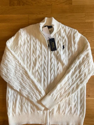 Vit kabelstickad tröja Polo Ralph Lauren - Kabelstickad vit tröja från Polo Ralph Lauren med dragkedja framtill och ribbade muddar. Tröjan har hög krage och den klassiska broderade loggan på bröstet. Perfekt för dig som gillar stilrena och tidlösa plagg.