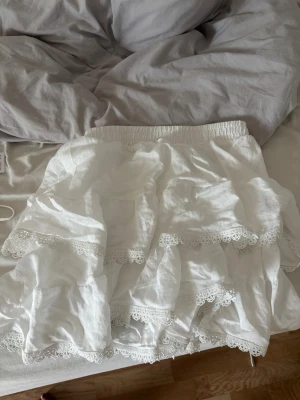 Vit volangkjol med spetsdetaljer - Söt vit kortkjol med flera lager volanger och fina spetsdetaljer längs kanterna. Kjolen har resår i midjan och är gjord i ett lätt material som passar perfekt till sommaren.