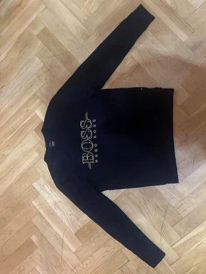 Svart sweatshirt från Hugo Boss - Stilren svart sweatshirt från Hugo Boss med stort guldfärgat BOSS-tryck på bröstet. Tröjan har rund halsringning och långa ärmar. Perfekt för en clean och modern look.