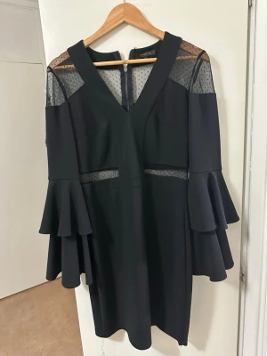Svart klänning med volangärm Mianotte - Svart kortklänning från Mianotte med v-ringning, volangärmar och transparenta partier i mesh med prickigt mönster. Klänningen har dragkedja bak och snygga detaljer som ger en unik look.