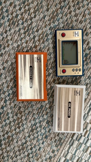 Nintendo Game & Watch konsoler - Tre klassiska Nintendo Game & Watch-konsoler: Donkey Kong (orange och silver), Oil Panic (vit och silver) och en enkel Game & Watch (blå och beige). Alla är i kompakt format med LCD-skärm och knappar i plast och metall. Perfekt för retrospelare och samlare. Dem kan nog fungera om man skaffar nya batterier.