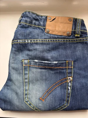 Dondup slim fit jeans - Snygga blå jeans med slitningar och coola kontrastsömmar i orange och gult. Klassisk femficksmodell med dekorativ bakficka och läderpatch i midjan. Perfekta för en avslappnad och trendig look.