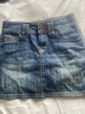 Blå jeanskjol med kontrastsömmar - Säljer en klassisk blå jeanskjol i kort modell med kontrastsömmar och röda detaljer vid linningen. Kjolen har fem fickor och bälteshällor, samt snygga dekorationer på bakfickorna. Perfekt till vardags för en avslappnad och trendig look.