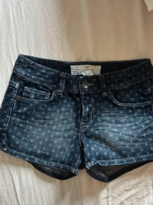 Lågmidjade shorts - Snygga mörkblå jeansshorts med diskret rutigt mönster och klassisk femficksdesign. Shortsen har låg midja, knappgylf och bälteshällor. Perfekta för sommaren och lätta att matcha med olika toppar.
