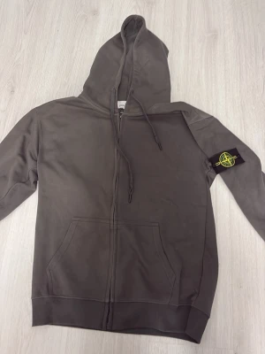Grå hoodie från Stone Island - Säljer en grå zip-hoodie från Stone Island med klassisk logopatch på vänster ärm. Hoodien har dragsko i huvan, känguruficka framtill och ribbade muddar. Tillverkad i mjukt bomullsmaterial som känns skönt mot huden. Perfekt för en avslappnad och stilren look. Startpris 750kr