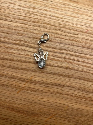 Lyckoängel i silver  - Söt berlock formad som en ängel med facetterade glaspärlor och silvriga metallvingar. Fästet är en karbinhake i metall, perfekt att hänga på väska, nyckelring eller smycke. Ger en glittrig och unik touch till din stil.