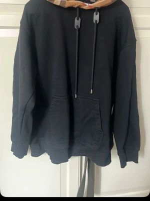 Svart hoodie med rutmönstrad huva - Snygg svart hoodie med stor ficka framtill och en huva i brunt rutmönster med dragkedjor. Hoodien har långa ärmar, ribbade muddar och justerbara snören i huvan. Perfekt för dig som gillar streetwear och vill sticka ut lite extra.