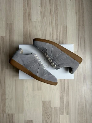 Grå sneakers från Maison Margiela - Snygga grå sneakers från Maison Margiela med ovandel i mocka och vita skosnören. Skorna har en klassisk siluett och en kontrasterande brun gummisula som ger en retrovibe. Perfekta för dig som gillar stilrena och tidlösa sneakers till vardagslooken.